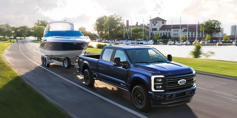 Blue Ford pickup truck towing a boat on a trailer along a lakeside road.