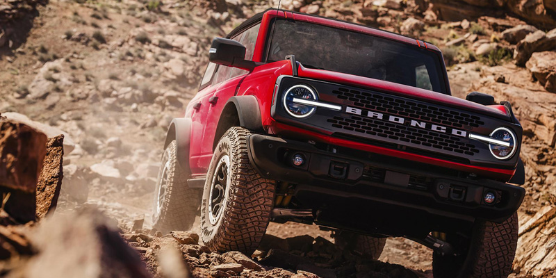 Red Ford Bronco driving through rocky off-road terrain kicking up dust among canyon rocks.